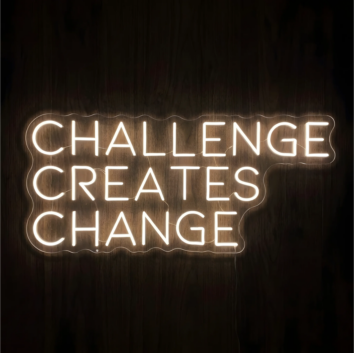 βCHALLENGE CREATES CHANGEβ LED Neon Sign glowing in warm white uppercase letters across three lines on clear acrylic against a dark wood background, suited to home offices and fitness spaces.