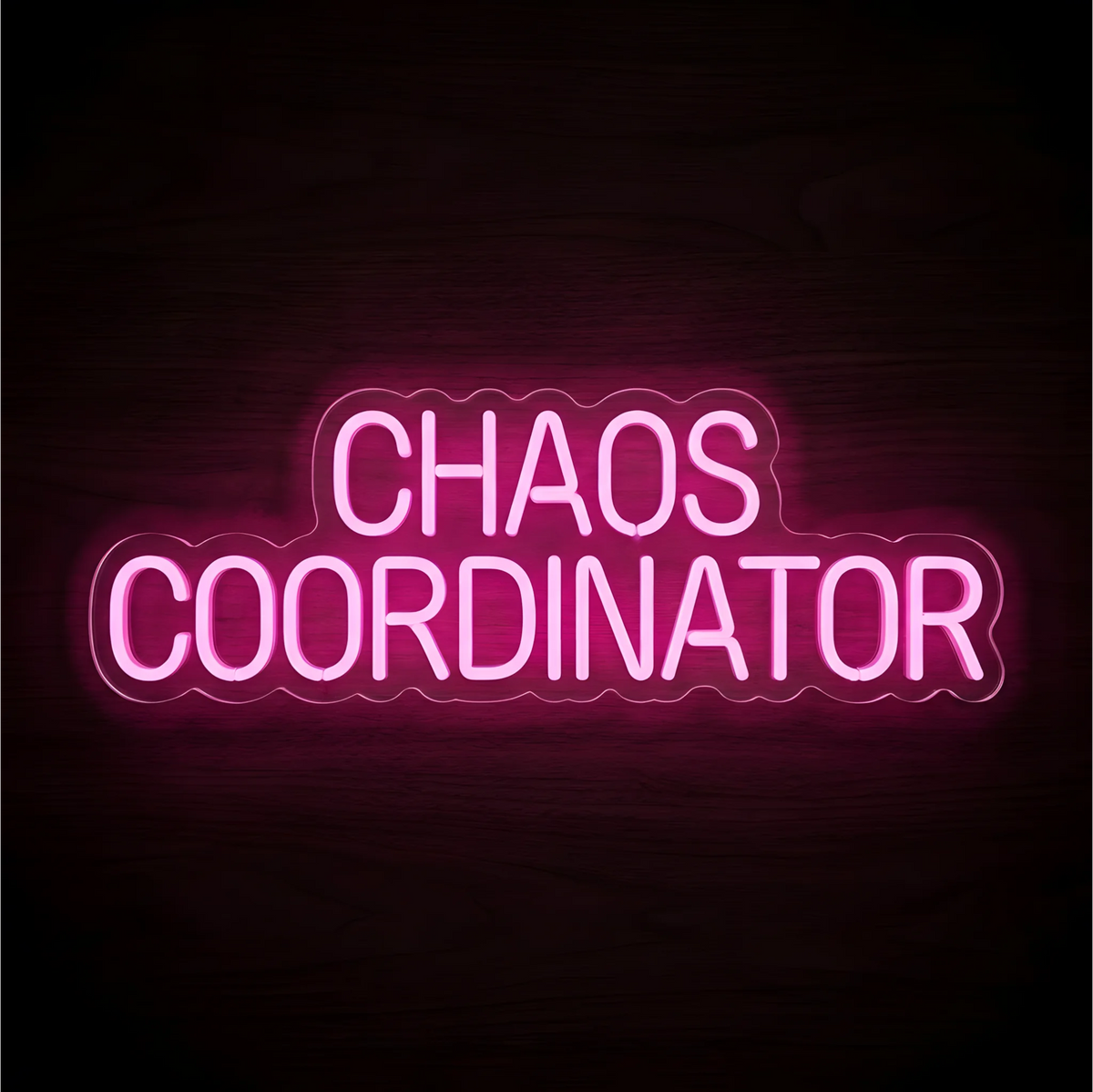 “CHAOS COORDINATOR” LED Neon Sign glowing in bright pink uppercase lettering across two lines on clear acrylic against a dark wood background, suited to home offices and team workspaces.