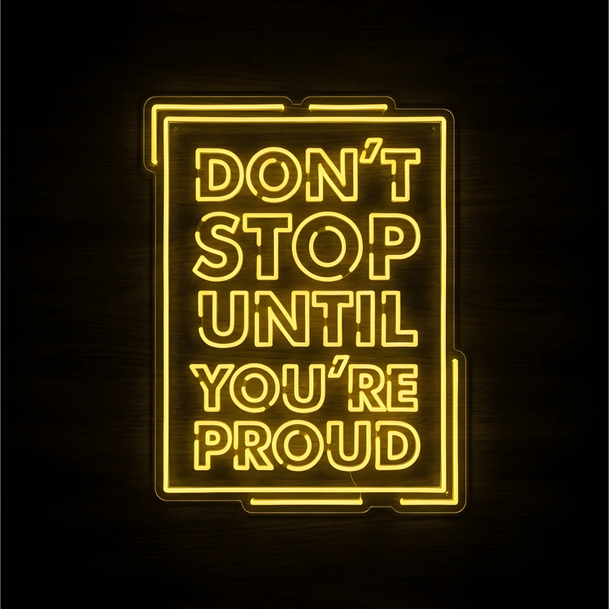 “DON’T STOP UNTIL YOU’RE PROUD” LED Neon Sign glowing yellow inside a rectangular neon outline on clear acrylic backing against a dark wood background, suited to home offices, gyms, and motivational walls.