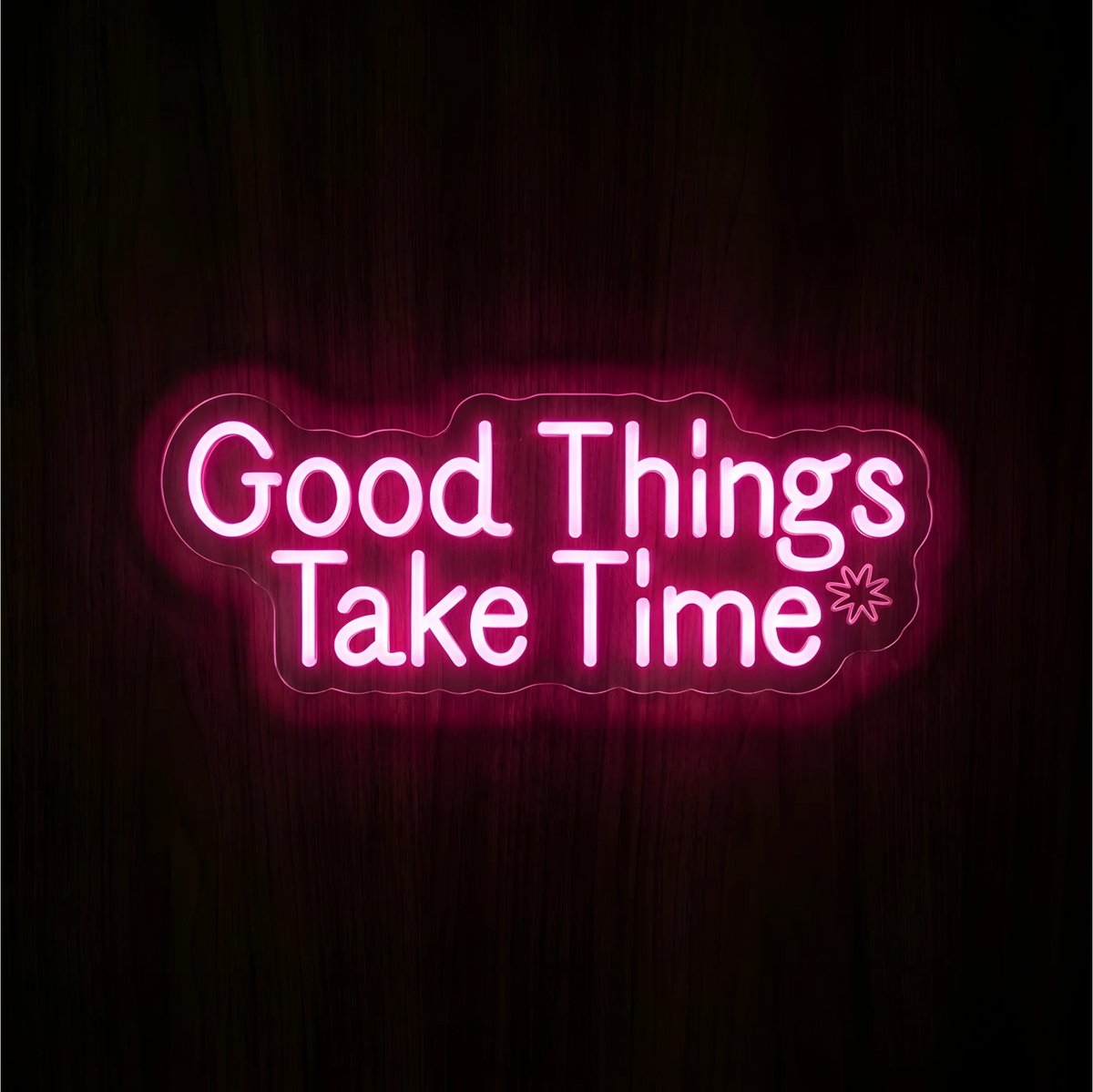 “Good Things Take Time” LED Neon Sign glowing in pink lettering with a small outlined starburst on clear acrylic against a dark wood background, suited to bedrooms and home offices.