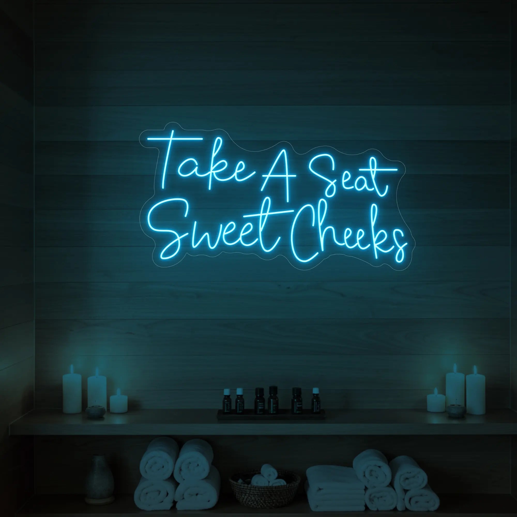 Photo of the 'Take A Seat Sweet Cheeks' neon sign glowing in ice blue against a styled interior wall.