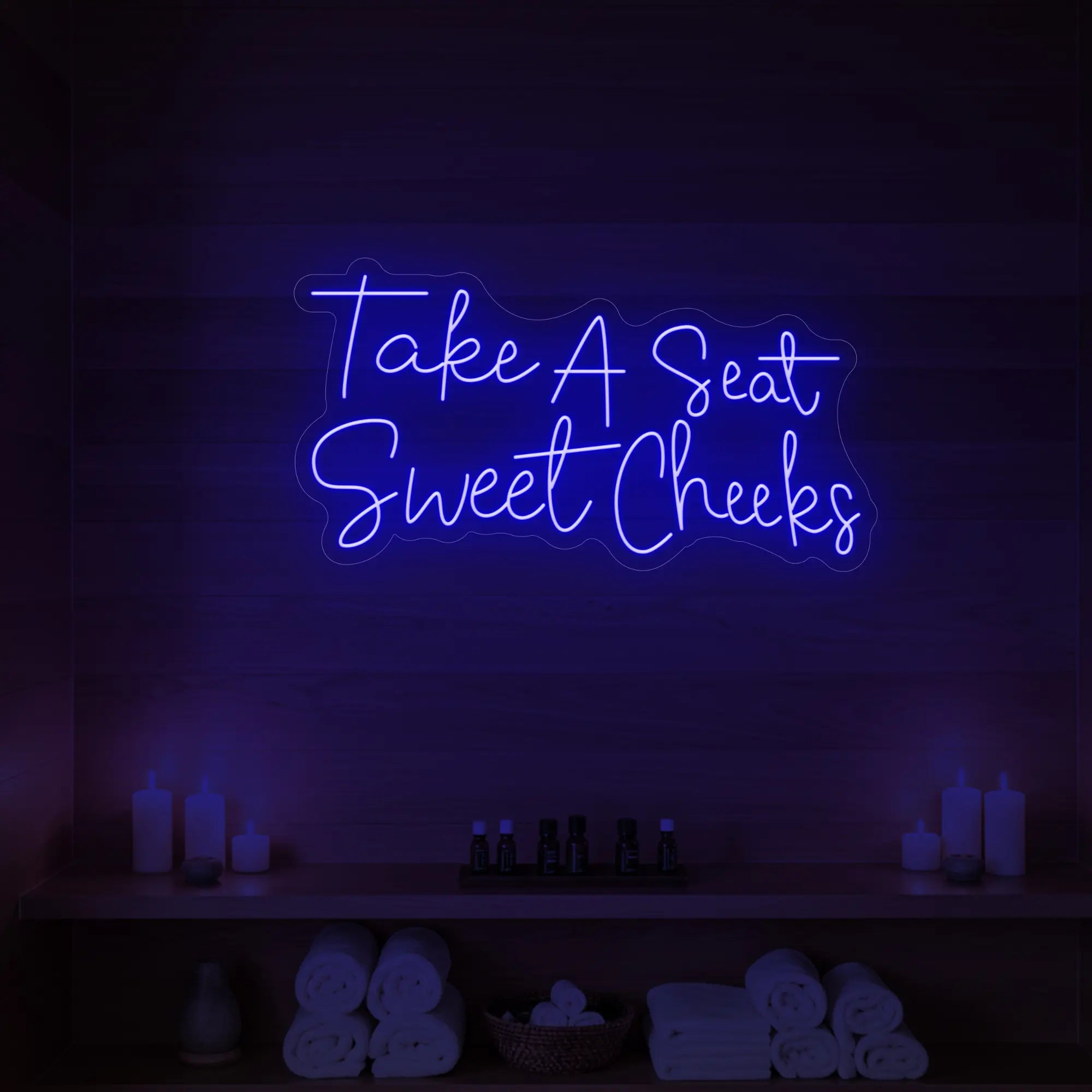Photo of the 'Take A Seat Sweet Cheeks' neon sign glowing in deep blue against a styled interior wall.