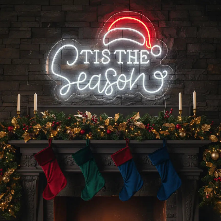 Photo of the 'Tis the Season with Santa Hat' neon sign glowing in cool white against a styled interior wall.