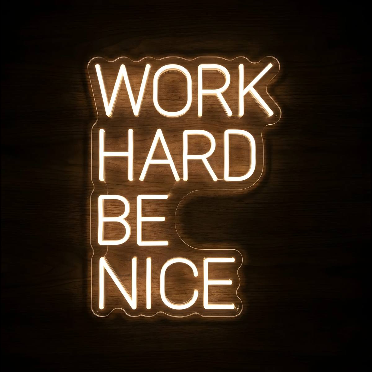 βWORK HARD BE NICEβ LED Neon Sign glowing in warm white lettering on clear acrylic backing against a dark wood background, suitable for office, classroom, and reception area decor.