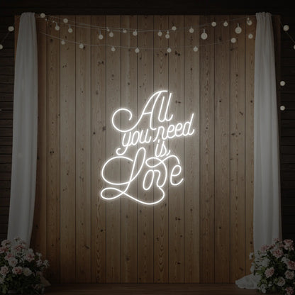 Photo of the 'All You Need Is Love V2' neon sign glowing in cool white against a styled interior wall.