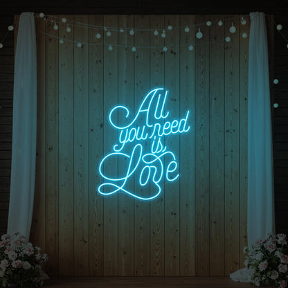 Photo of the 'All You Need Is Love V2' neon sign glowing in ice blue against a styled interior wall.