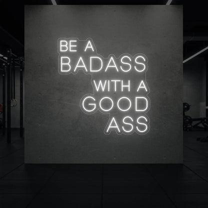 'Be a Badass With a Good Ass' neon sign glowing in cool white against a styled interior wall.