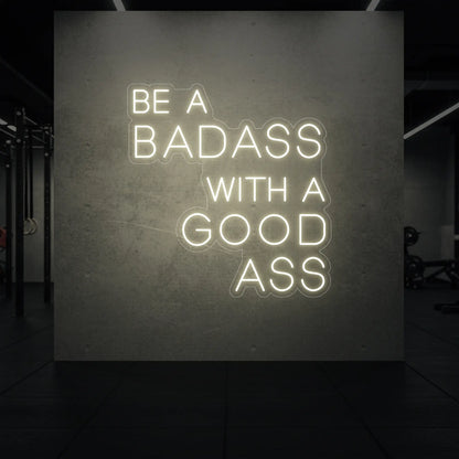  'Be a Badass With a Good Ass' neon sign glowing in warm white against a styled interior wall.