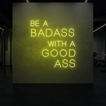 'Be a Badass With a Good Ass' neon sign glowing in lemon yellow against a styled interior wall.
