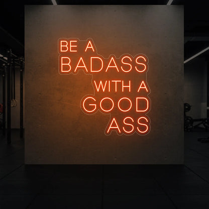  'Be a Badass With a Good Ass' neon sign glowing in golden yellow against a styled interior wall.