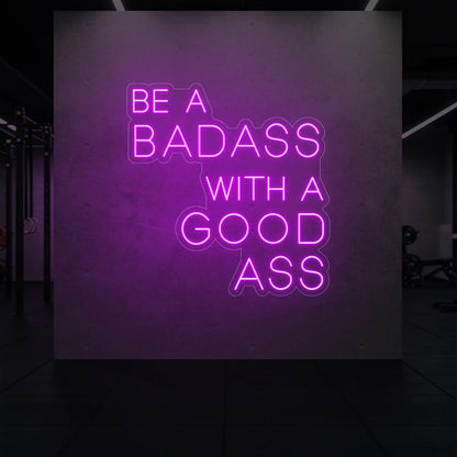'Be a Badass With a Good Ass' neon sign glowing in deep pink against a styled interior wall.