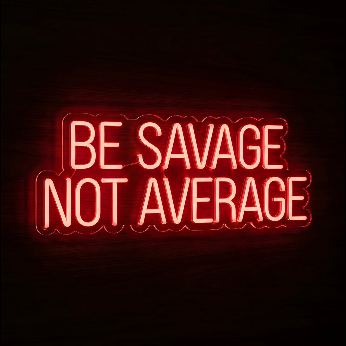 “BE SAVAGE NOT AVERAGE” LED Neon Sign glowing in bright red uppercase lettering across two lines on clear acrylic against a dark wood background, suited to home offices and gym spaces.