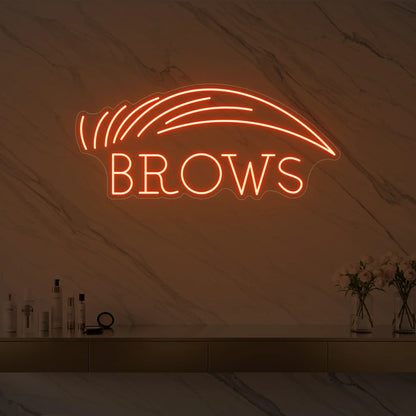 Photo of the 'BROWS' neon sign with an eyebrow graphic glowing in golden yellow against a styled interior wall.