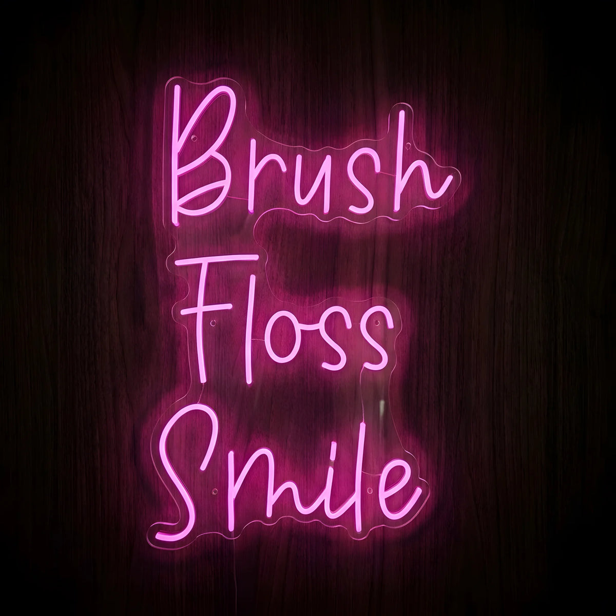 Brush Floss Smile LED Neon Sign featuring bright pink cursive lettering arranged vertically on clear acrylic backing against a dark wooden wall, perfect for dental office decor.