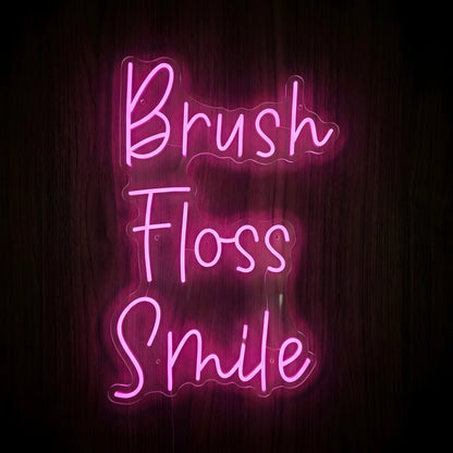 Brush Floss Smile LED Neon Sign featuring bright pink cursive lettering arranged vertically on clear acrylic backing against a dark wooden wall, perfect for dental office decor.