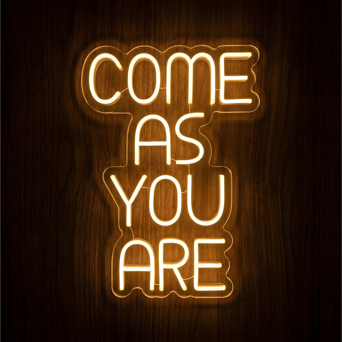 “COME AS YOU ARE” LED Neon Sign with warm white lettering on clear acrylic backing, glowing against a dark wood background and suited to living rooms, entryways, or reception areas.