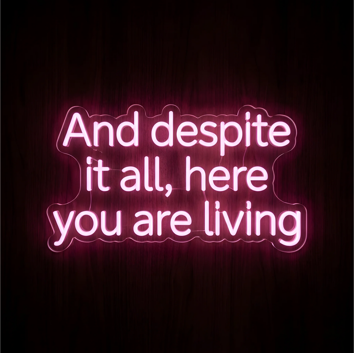 “And despite it all, here you are living” LED Neon Sign in glowing pink lettering on clear acrylic against a dark wood background, ideal for bedrooms and inspirational wall decor.