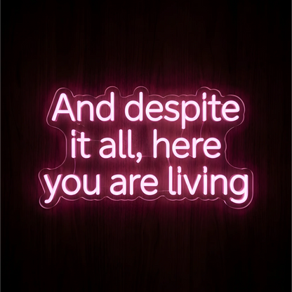 “And despite it all, here you are living” LED Neon Sign in glowing pink lettering on clear acrylic against a dark wood background, ideal for bedrooms and inspirational wall decor.