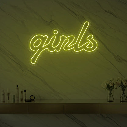 Photo of the 'girls' neon sign in playful script glowing in lemon yellow against a styled interior wall.