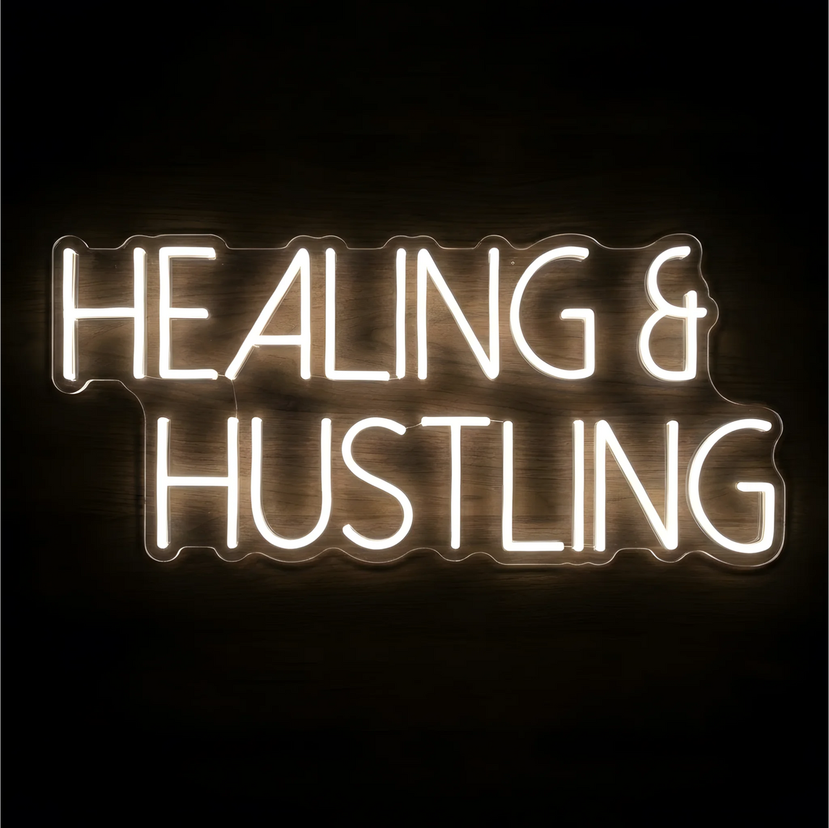“Healing & Hustling” LED Neon Sign glowing in bright white uppercase lettering on clear acrylic against a dark wood background, suited to home offices and creative workspaces.