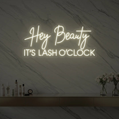 Photo of the 'Hey Beauty IT’S LASH O’CLOCK' neon sign glowing in warm white against a styled interior wall.