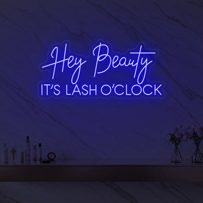 Photo of the 'Hey Beauty IT’S LASH O’CLOCK' neon sign glowing in deep blue against a styled interior wall.
