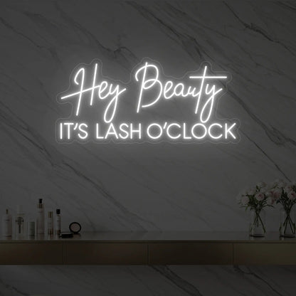 Photo of the 'Hey Beauty IT’S LASH O’CLOCK' neon sign glowing in cool white against a styled interior wall.
