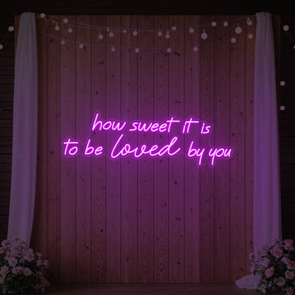 Photo of the 'How Sweet It Is To Be Loved By You' neon sign glowing in deep pink against a styled interior wall.