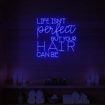 Photo of the 'Life isn’t perfect but your hair can be' neon sign glowing in deep blue against a wood-paneled spa wall.