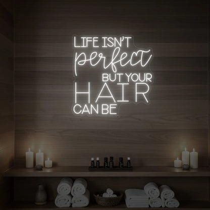 Photo of the 'Life isn’t perfect but your hair can be' neon sign glowing in cool white against a wood-paneled spa wall.