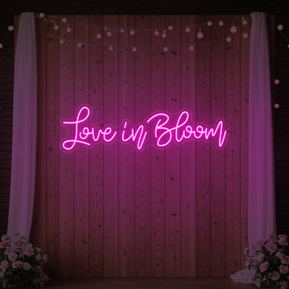 Photo of the 'Love in Bloom' neon sign glowing in light pink against a styled interior wall.