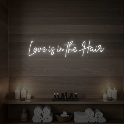 Photo of the 'Love is in the Hair' neon sign glowing in cool white against a styled interior wall.