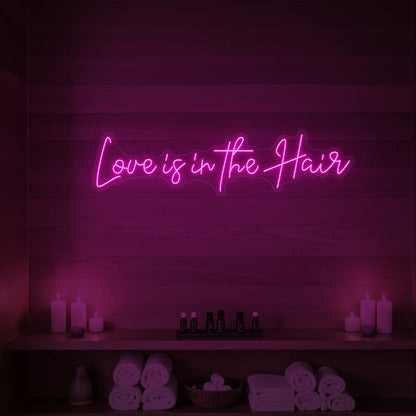 Photo of the 'Love is in the Hair' neon sign glowing in light pink against a styled interior wall.