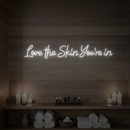 Photo of the 'Love the Skin You're In' neon sign glowing in cool white against a styled interior wall.