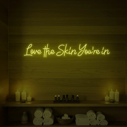 Photo of the 'Love the Skin You're In' neon sign glowing in lemon yellow against a styled interior wall.
