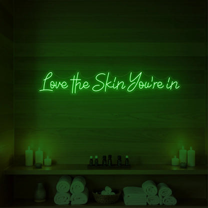 Photo of the 'Love the Skin You're In' neon sign glowing in green against a styled interior wall.