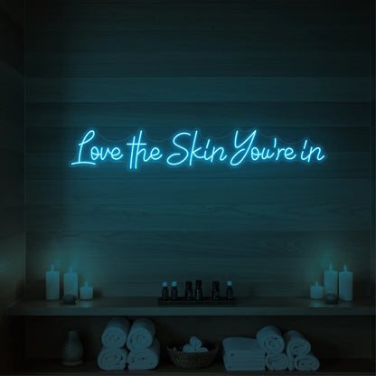 Photo of the 'Love the Skin You're In' neon sign glowing in ice blue against a styled interior wall.