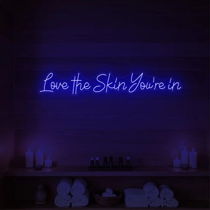 Photo of the 'Love the Skin You're In' neon sign glowing in deep blue against a styled interior wall.