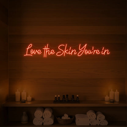 Photo of the 'Love the Skin You're In' neon sign glowing in golden yellow against a styled interior wall.