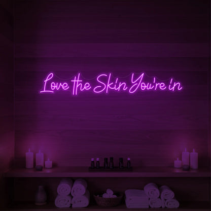 Photo of the 'Love the Skin You're In' neon sign glowing in deep pink against a styled interior wall.