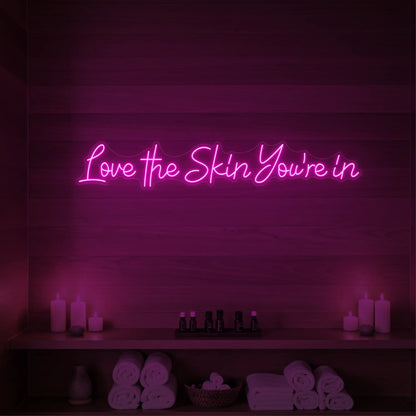 Photo of the 'Love the Skin You're In' neon sign glowing in light pink against a styled interior wall.