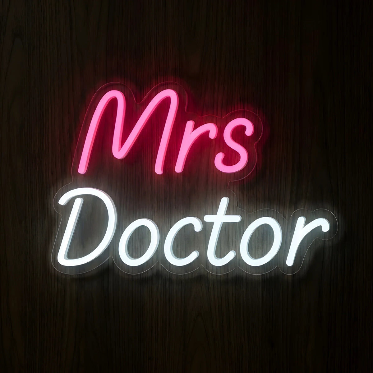 Mrs Doctor Script LED Neon Sign featuring glowing pink Mrs lettering above elegant white Doctor cursive text on clear cut-to-shape acrylic backing against a dark wooden wall.