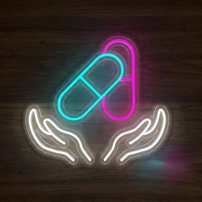 Pharmacy Pill Care LED Neon Sign featuring glowing cyan and pink capsules above two white caring hands on clear cut-to-shape acrylic against a dark wooden wall.