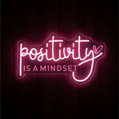 Positivity Is a Mindset LED Neon Sign in pink and white lettering with a small heart at the end of the script, displayed on clear acrylic against a dark wood background, suited to bedrooms and wellness spaces.