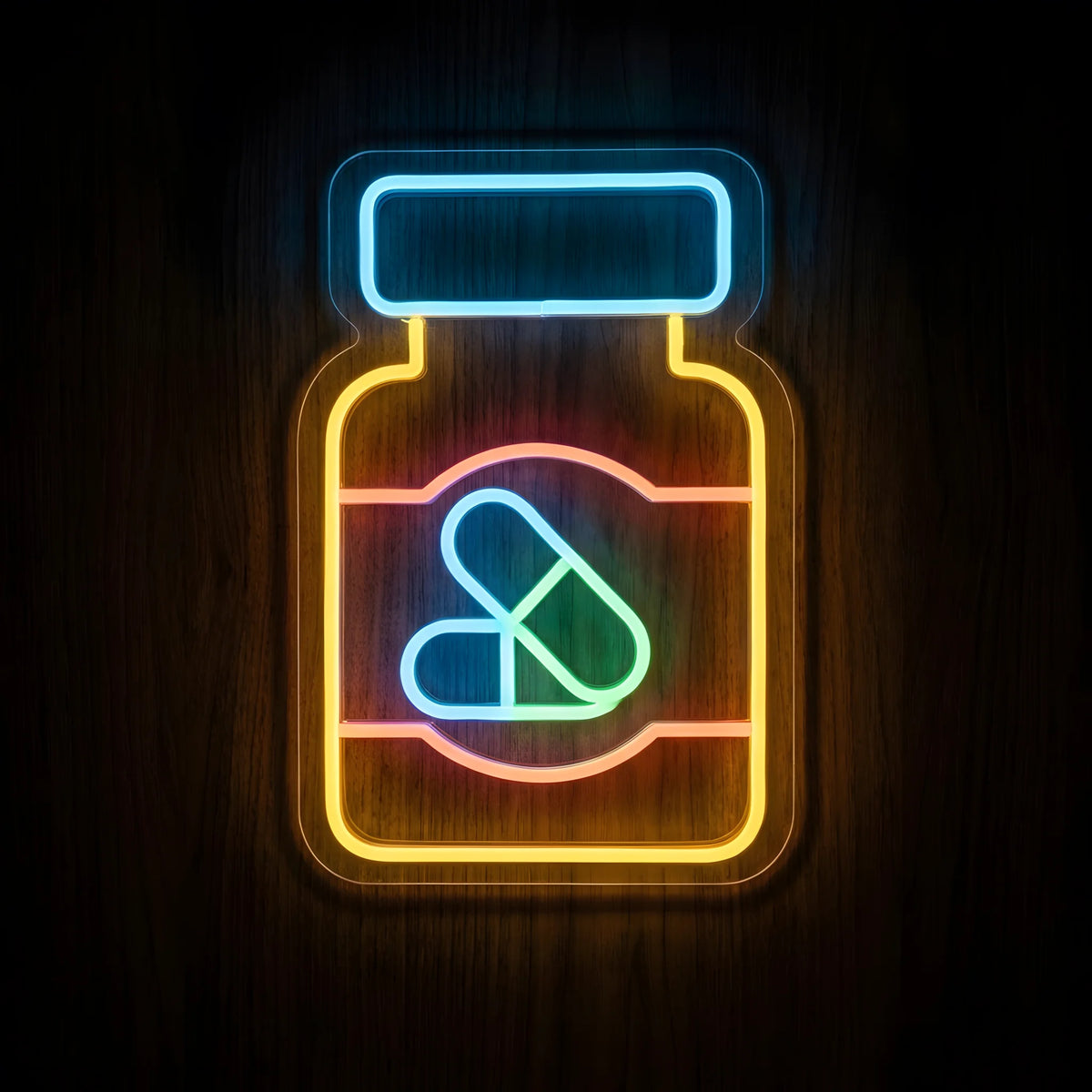 Prescription Pill Bottle LED Neon Sign featuring a yellow medicine bottle, blue cap, pink label, and blue and green capsules on clear acrylic backing against a dark wooden wall.
