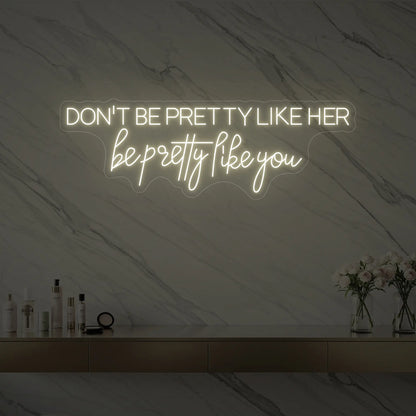 Photo of the 'DON'T BE PRETTY LIKE HER — be pretty like you' neon sign glowing in warm white against a styled interior wall.