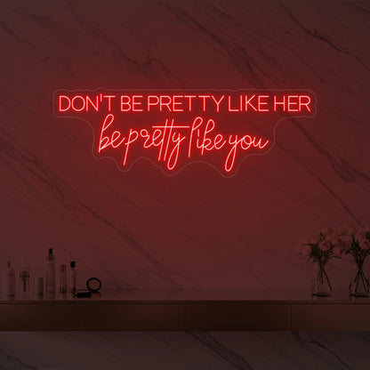 Photo of the 'DON'T BE PRETTY LIKE HER — be pretty like you' neon sign glowing in red against a styled interior wall.