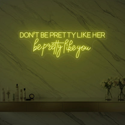 Photo of the 'DON'T BE PRETTY LIKE HER — be pretty like you' neon sign glowing in lemon yellow against a styled interior wall.
