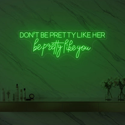 Photo of the 'DON'T BE PRETTY LIKE HER — be pretty like you' neon sign glowing in green against a styled interior wall.