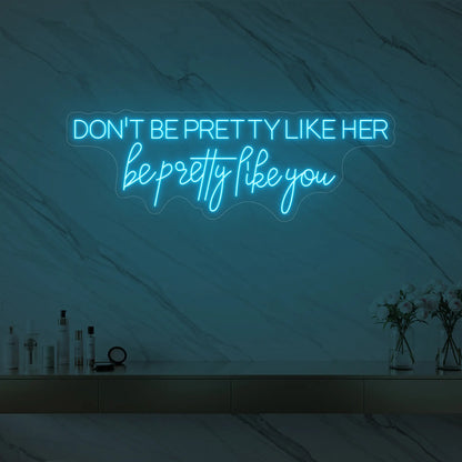 Photo of the 'DON'T BE PRETTY LIKE HER — be pretty like you' neon sign glowing in ice blue against a styled interior wall.
